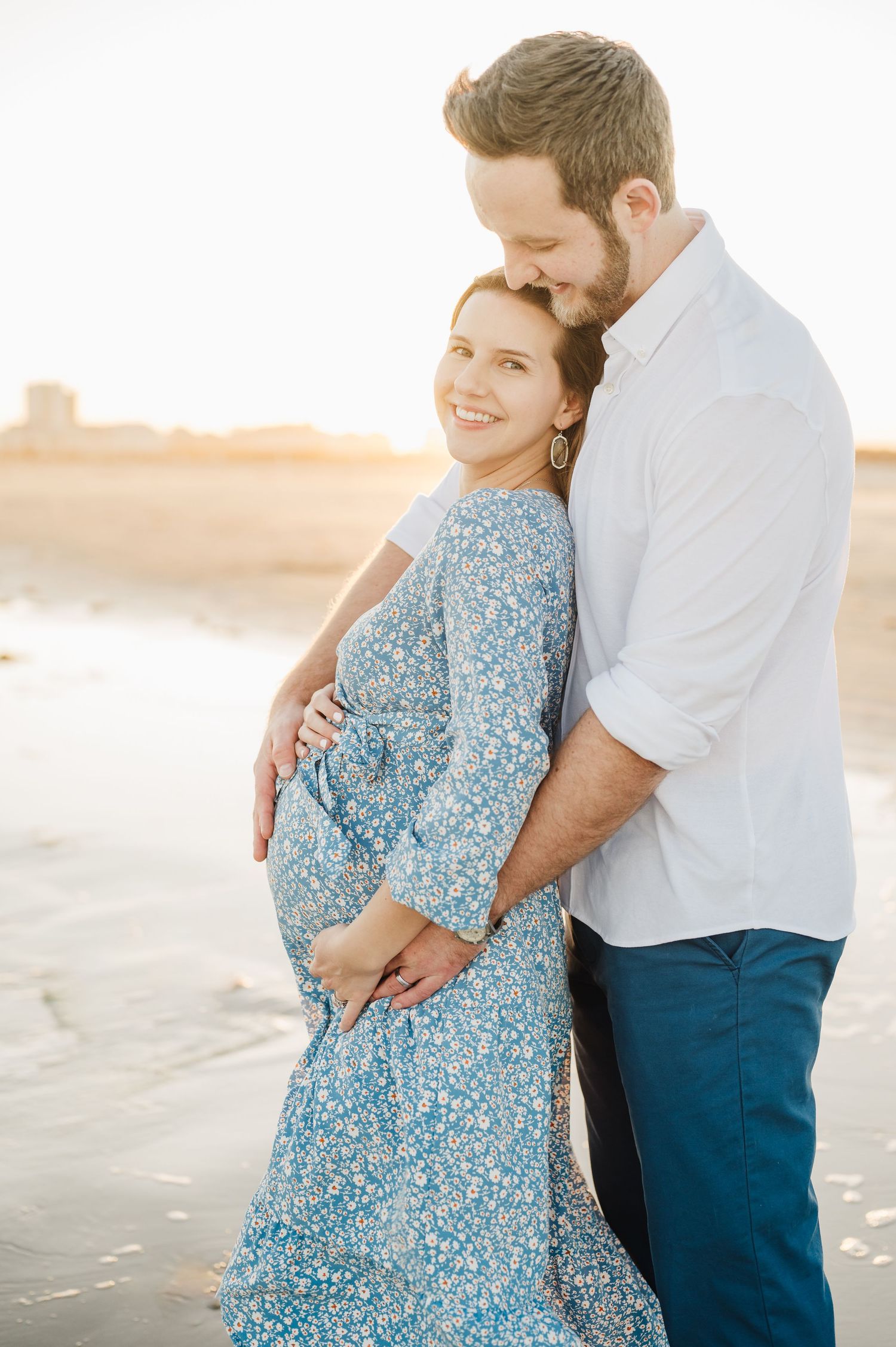 A couple embraces on a beach at sunset wearing blue and white, creating a romantic coastal portrait series.