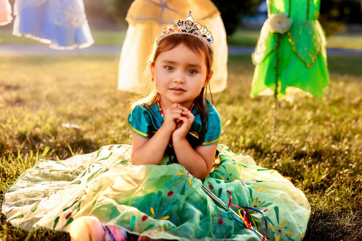 A young girl in a green princess dress and tiara sits on grass at an outdoor party.