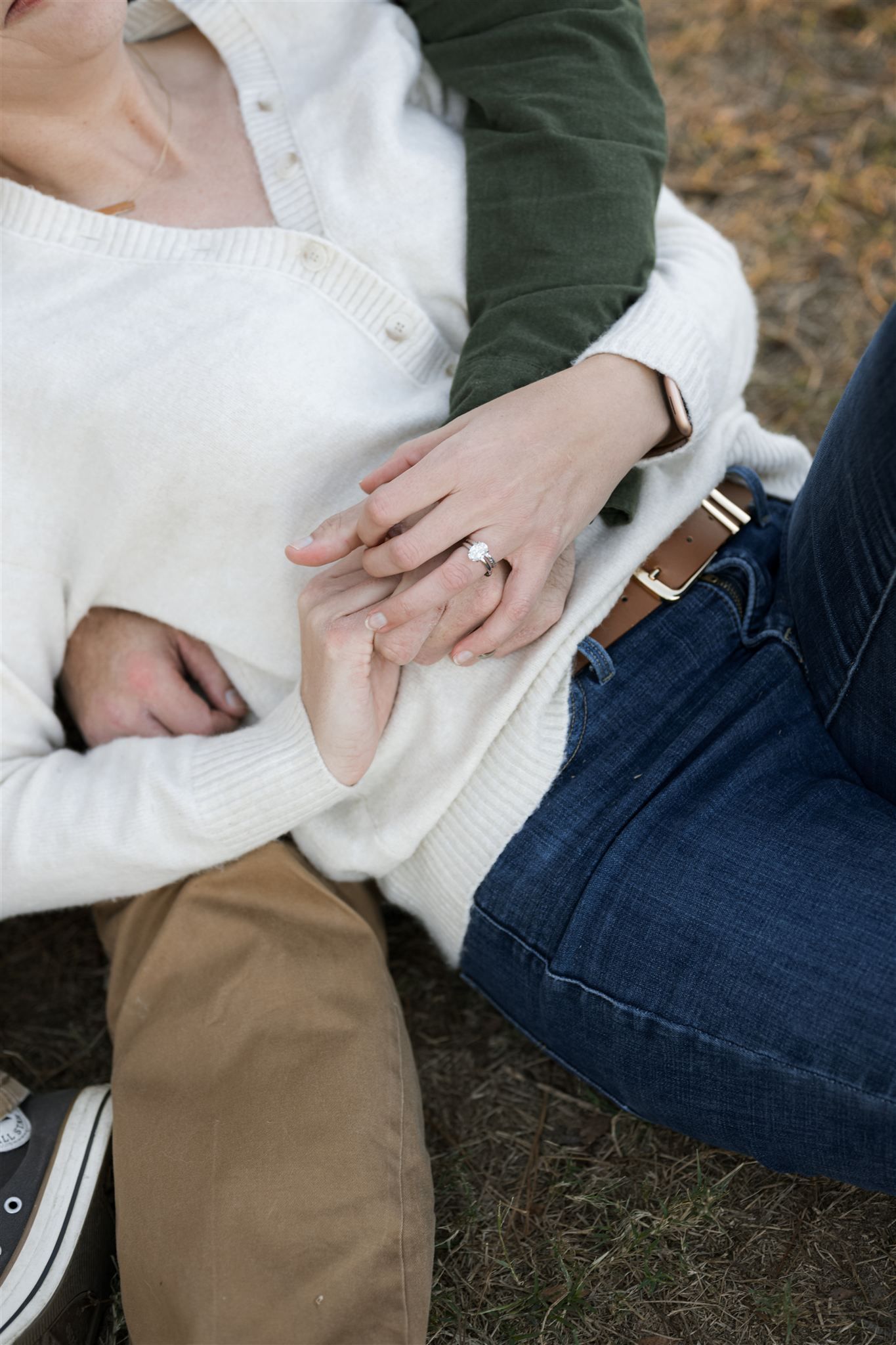 Close-up of couple holding hands outdoors, woman wearing engagement ring, dressed in white sweater and jeans.