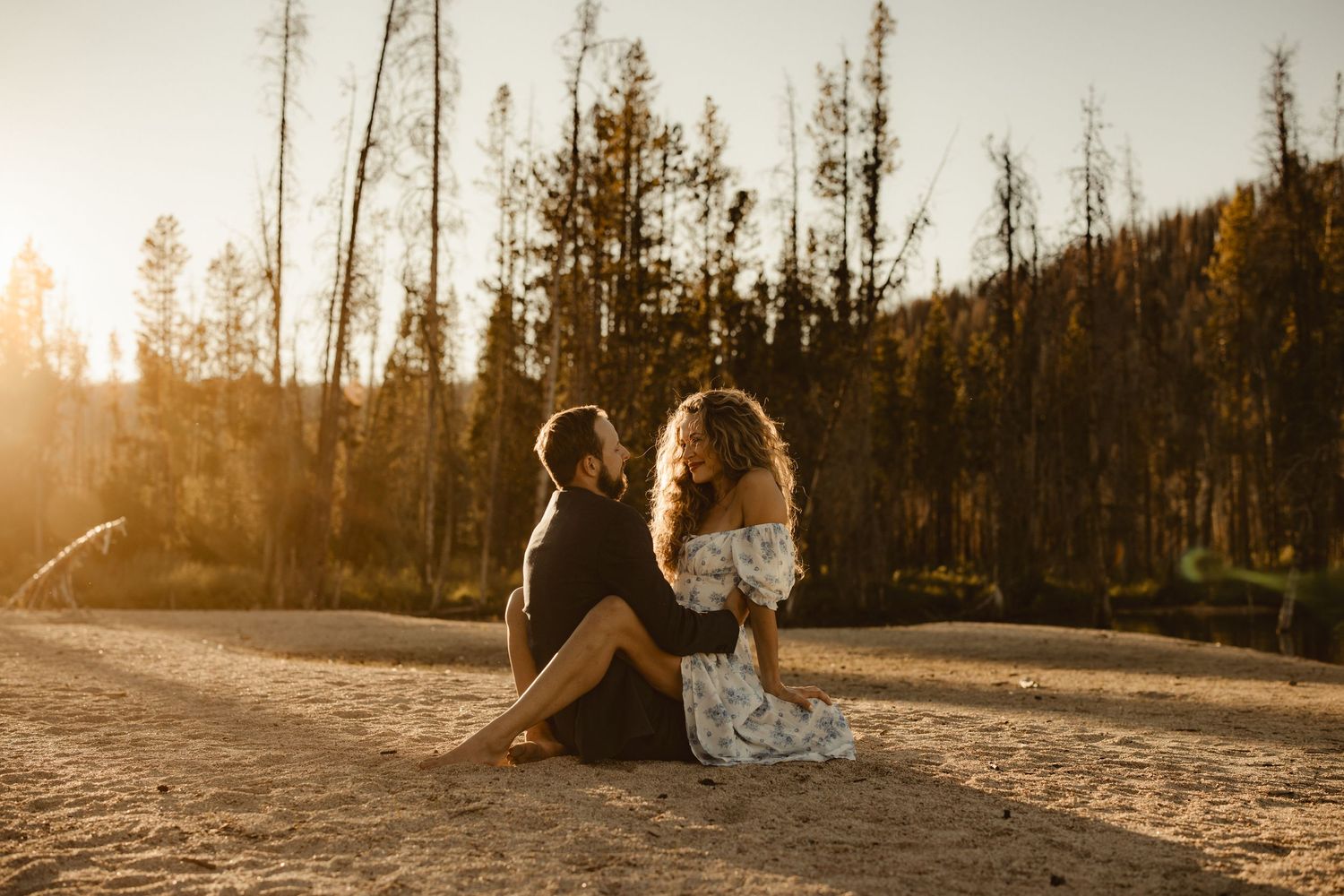 Silhouettes of a couple backlit by golden sunlight among tall trees at sunset.