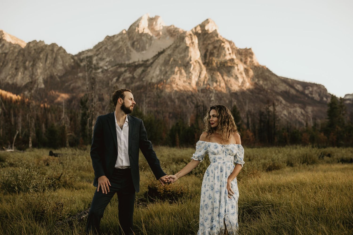 A couple walks hand in hand through a meadow with dramatic mountain peaks in the background.