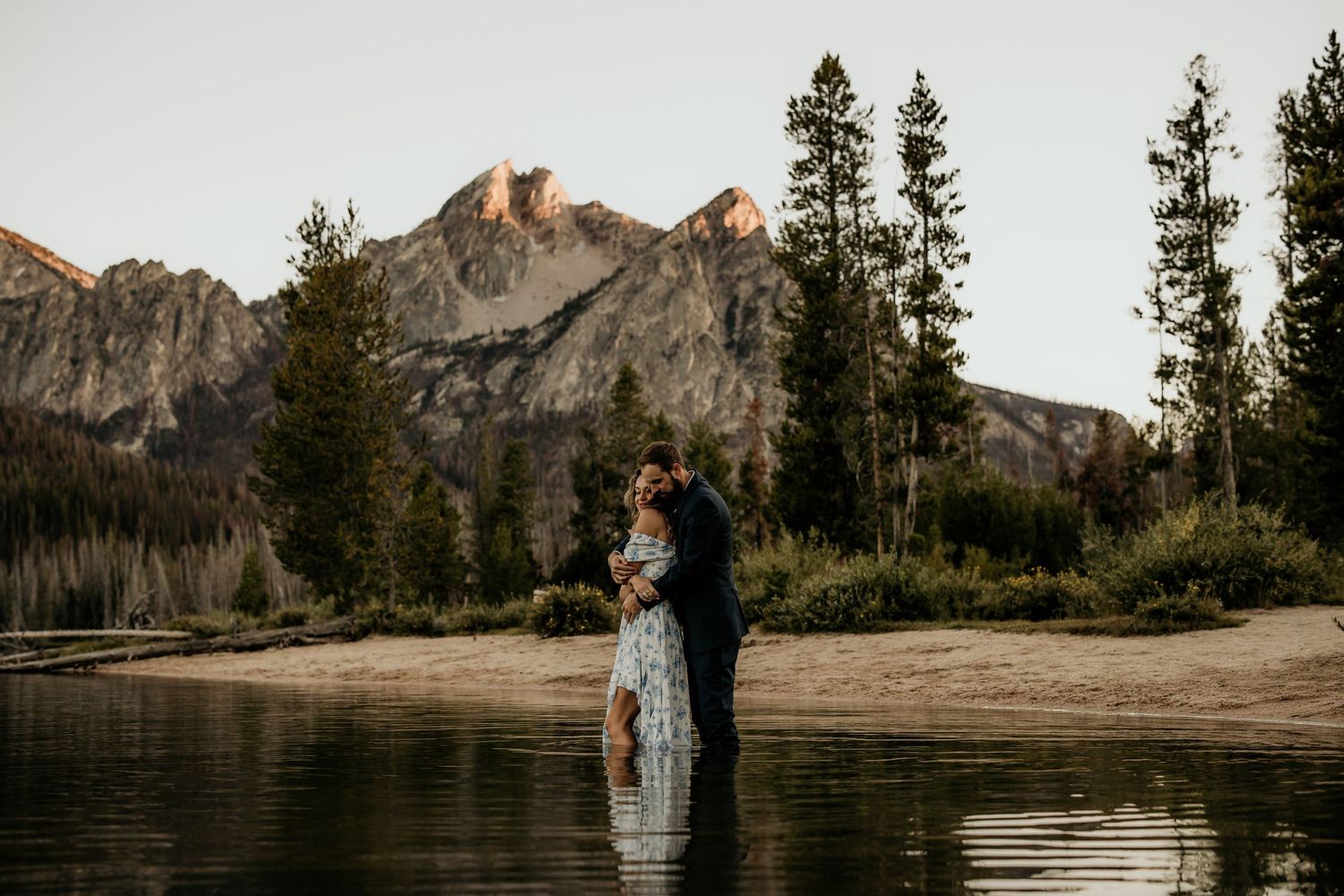 A couple stands in shallow water with mountain peaks and evergreen trees creating a dramatic backdrop at sunset.