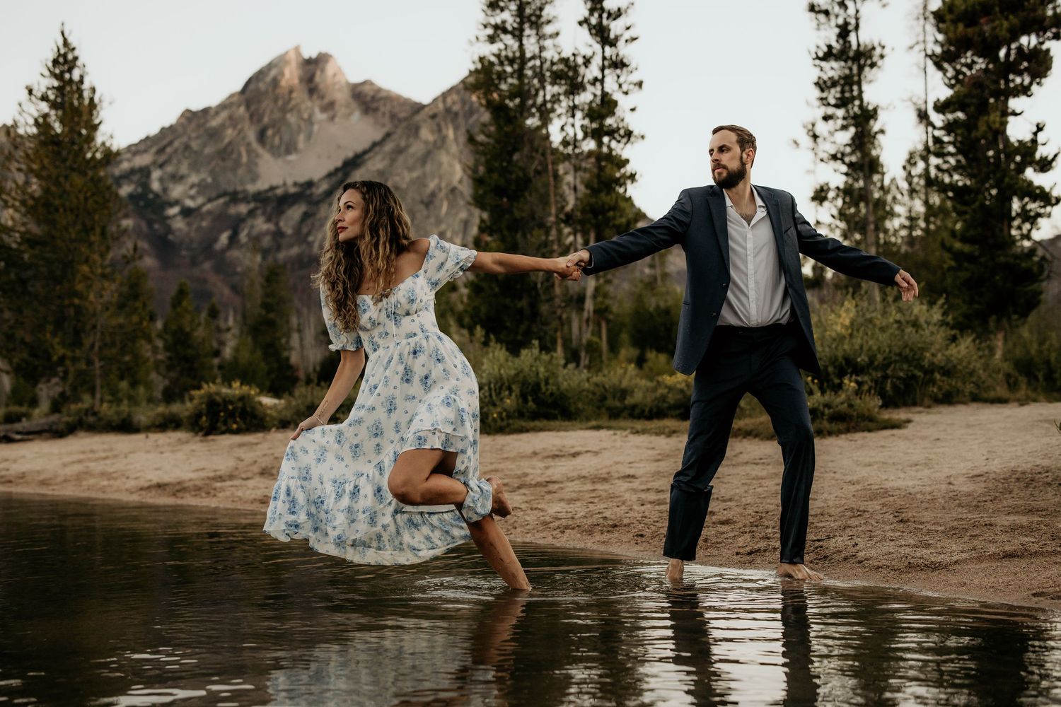 A couple dances playfully at the edge of a mountain lake surrounded by evergreen trees.