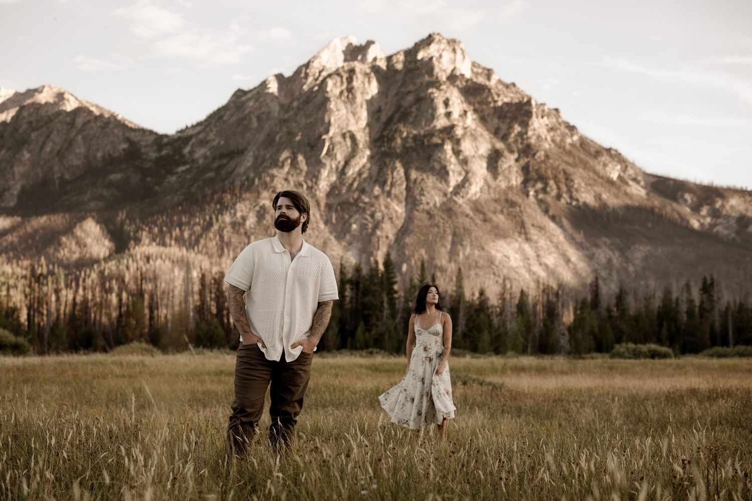 Couple poses in meadow with majestic mountain peaks in background.