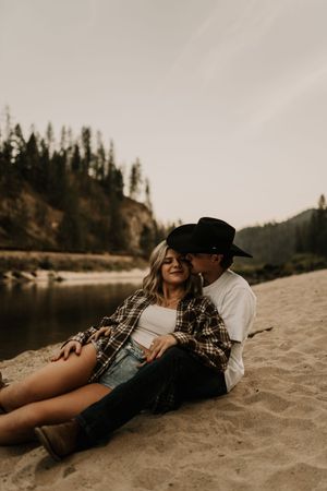 A couple relaxes together on a sandy beach near a forested lake shore.
