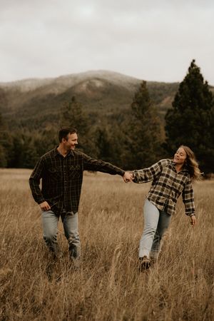 A playful couple dances through a golden mountain meadow with evergreen trees in the background.