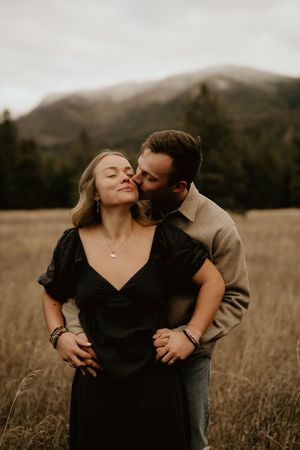 Couple shares intimate moment in golden field with mountains in background.