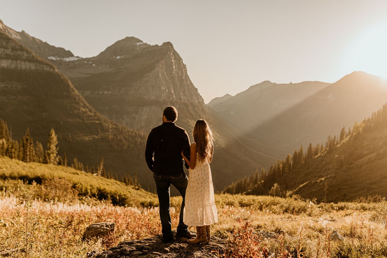A couple shares an intimate moment on a mountain trail during sunset with peaks in the distance.