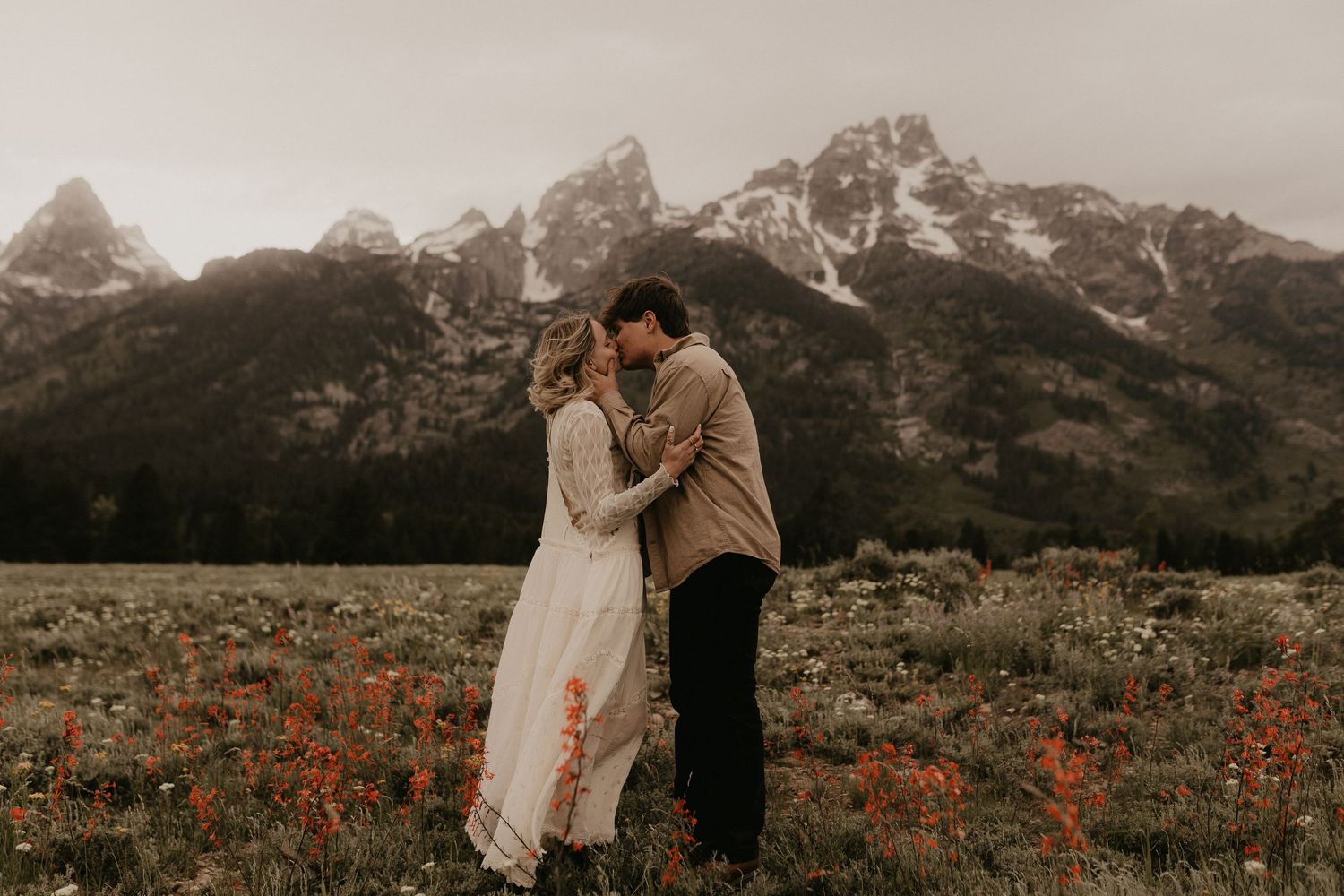 A romantic couple embraces in a wildflower meadow with majestic mountain peaks in the background at sunset.