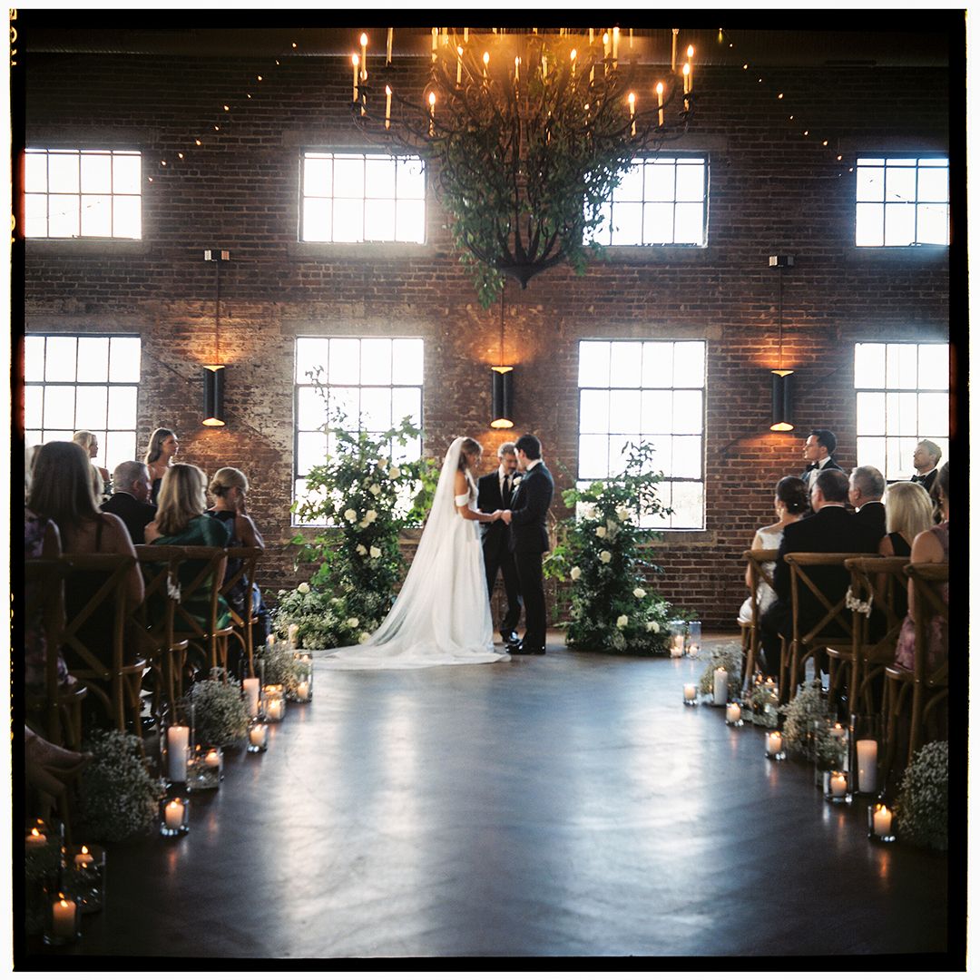 Wedding ceremony in a rustic brick-walled venue with natural light streaming through large windows and guests seated.
