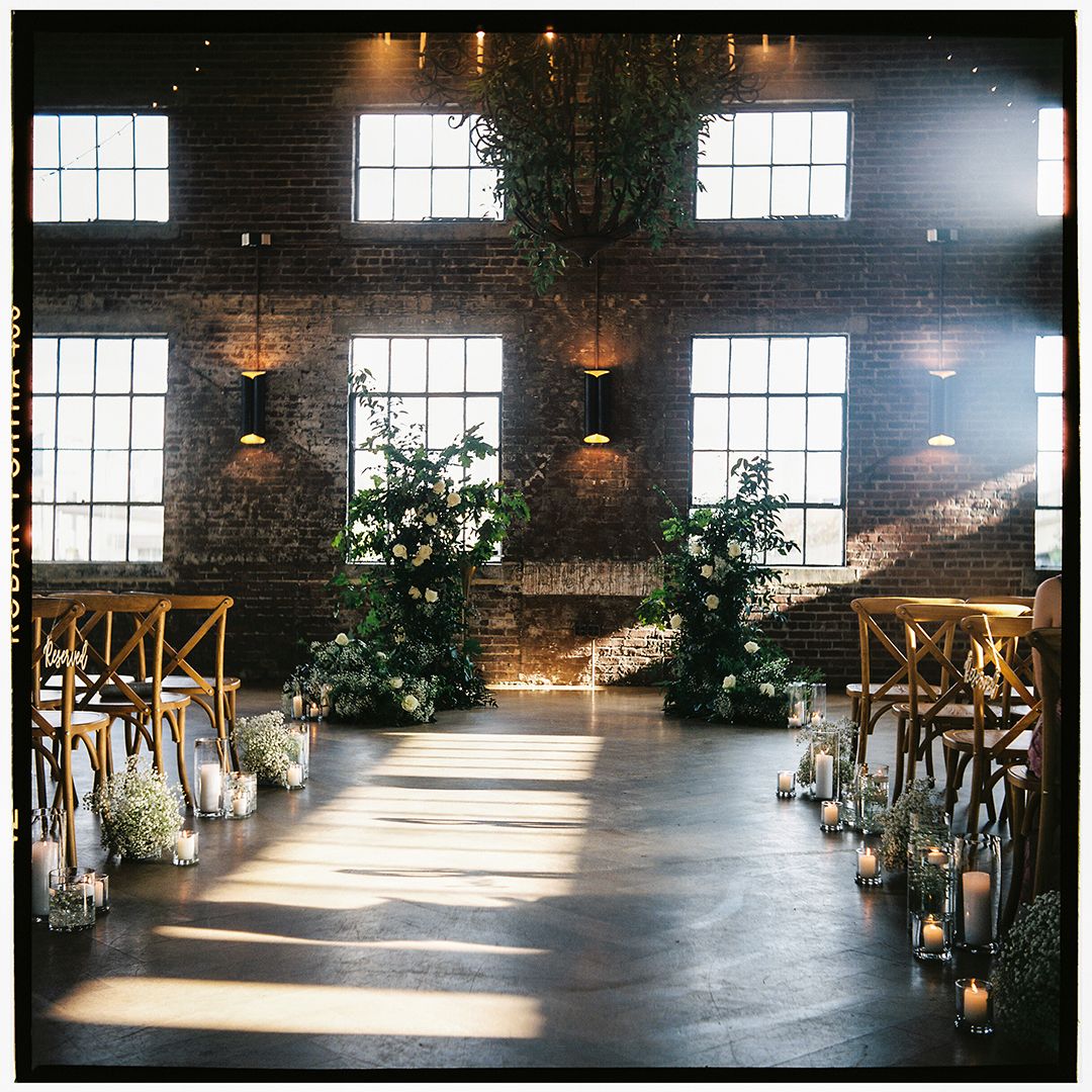 Sunlight streams through industrial windows in a rustic brick venue space with hardwood floors and hanging lights.