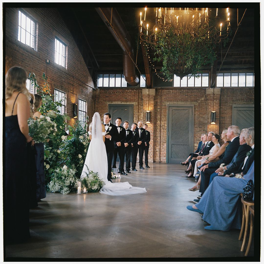 Indoor wedding ceremony in a rustic barn venue with elegant chandeliers and large windows illuminating the space.