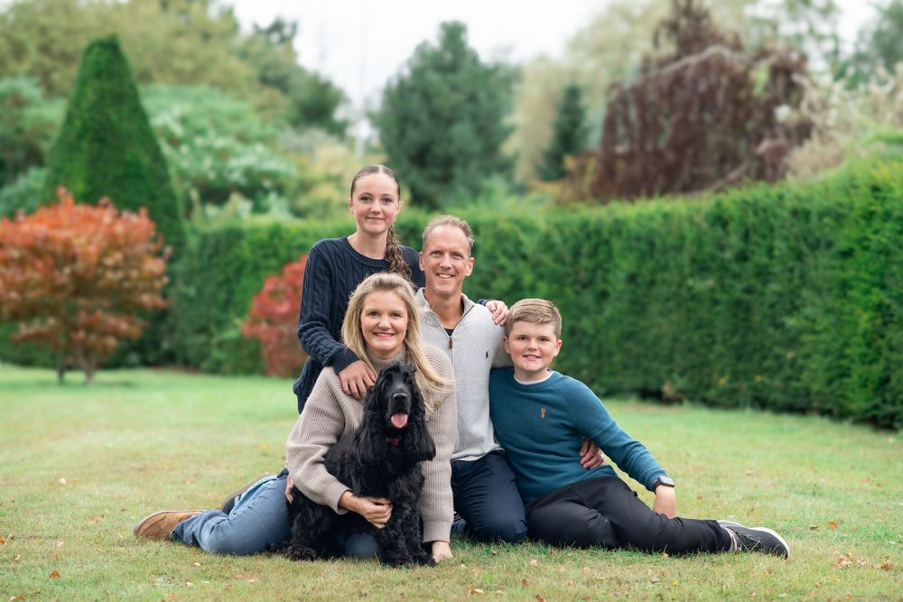 A family sits together with their on grass in a garden with tall hedges and colorful autumn foliage in the background in Leeds.