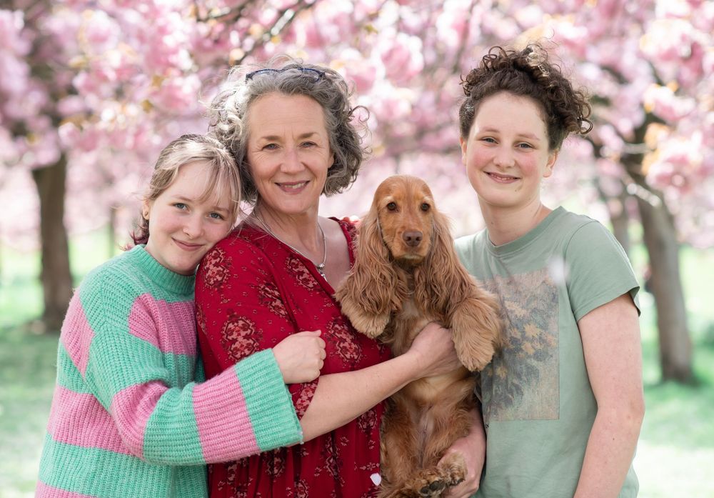 A mother and two teenage daughters stand together in front of the cherry blossom trees in Harrogate with their spaniel dog.
