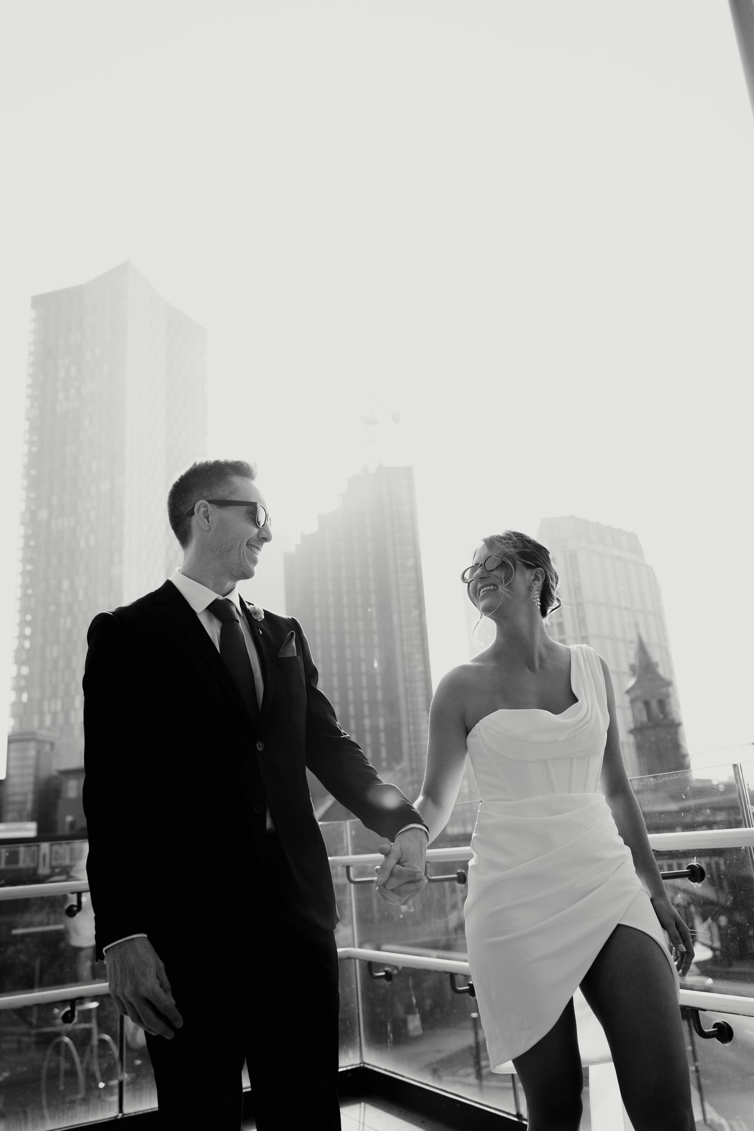 Two people in formal attire engage in conversation on a balcony with a cityscape backdrop in black and white.