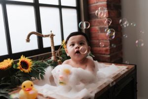 Baby in bubble bath plays near window with rubber duck and sunflowers.寶寶在泡泡浴裡玩耍，窗邊有橡皮鴨和向日葵。