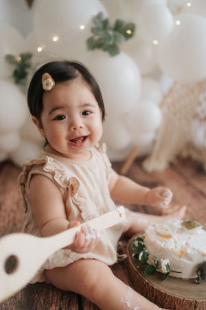 Baby in white lace dress sits on wooden surface surrounded by soft white decor and greenery.身穿白色蕾絲連身裙的嬰兒坐在木質表面上，周圍環繞著柔和的白色裝飾和綠植。