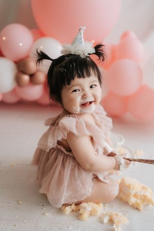 Baby in pink dress smiling during cake smash photo session with balloon backdrop.身穿粉紅色洋裝的寶寶在氣球背景下拍攝蛋糕砸臉照時笑容燦爛。