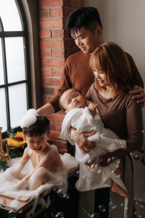 Family enjoying tender moment with children by brick window with floating bubbles.一家人依偎在磚窗邊，孩子們享受著漂浮泡泡的溫馨時刻。