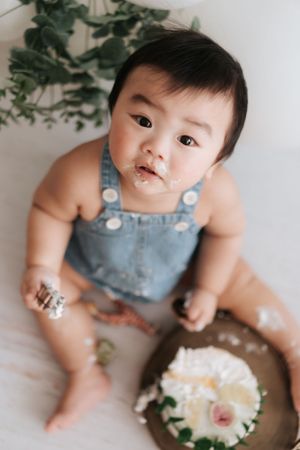 Baby in denim overalls sits with messy cake during first birthday celebration.寶寶穿著牛仔背帶褲，在一歲生日慶祝活動中，面前擺放著一塊弄得亂七八糟的蛋糕。
