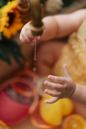 Baby hands reaching toward a dripping brass faucet with sunflowers and citrus fruits in the background.嬰兒的小手伸向滴水的黃銅水龍頭，背景是向日葵和柑橘類水果。