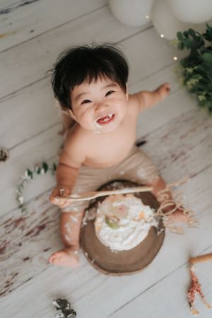 Happy baby enjoying cake smash session on rustic white wooden floor.快樂的寶寶在質樸的白色木地板上享受著蛋糕砸臉的樂趣。