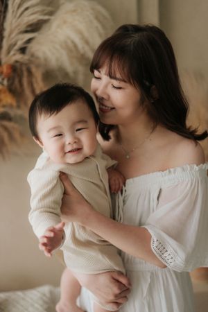 Mother in white dress holding smiling baby in beige outfit against neutral background身穿白色洋裝的母親抱著穿著米色衣服的微笑嬰兒，背景為中性色調。