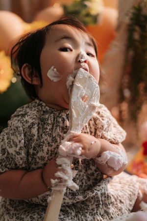 Baby in floral dress messily eating whipped cream from a mixer beater with cream on their cheeks.穿著碎花裙的寶寶狼吞虎嚥地吃著攪拌器打發的奶油，臉頰上沾滿了奶油。