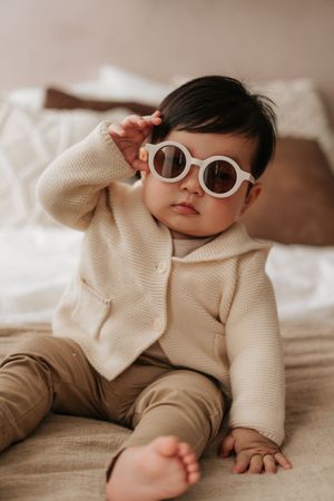 Baby wearing knit cream sweater and retro white sunglasses lies on soft beige bedding in indoor lifestyle photoshoot.身穿奶油色針織毛衣、戴著復古白色太陽眼鏡的嬰兒