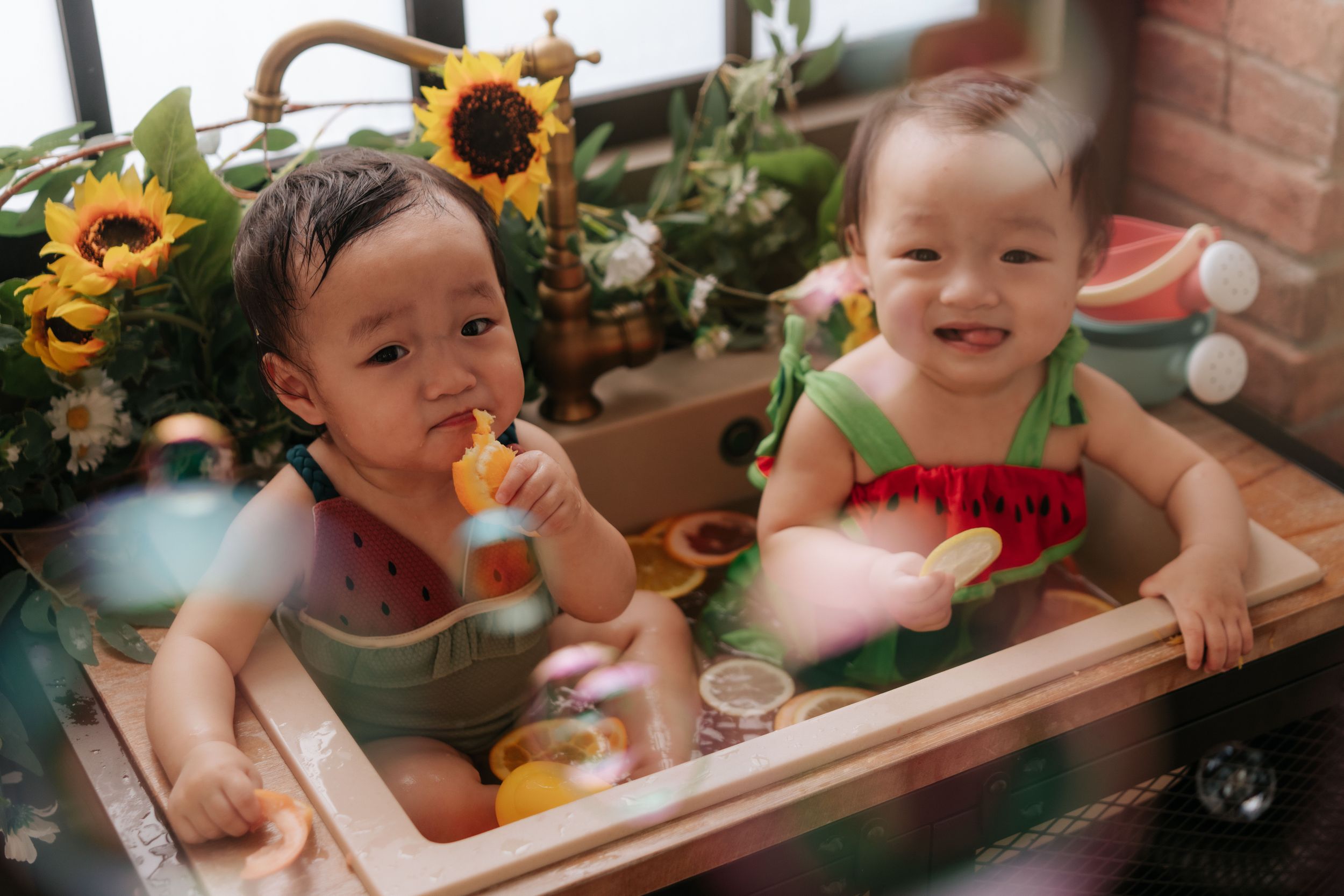 Two smiling toddlers in watermelon outfits sitting in a wooden tub surrounded by sunflowers and green plants.兩個穿著西瓜圖案衣服的笑容燦爛的幼兒坐在一個木製浴缸裡，周圍環