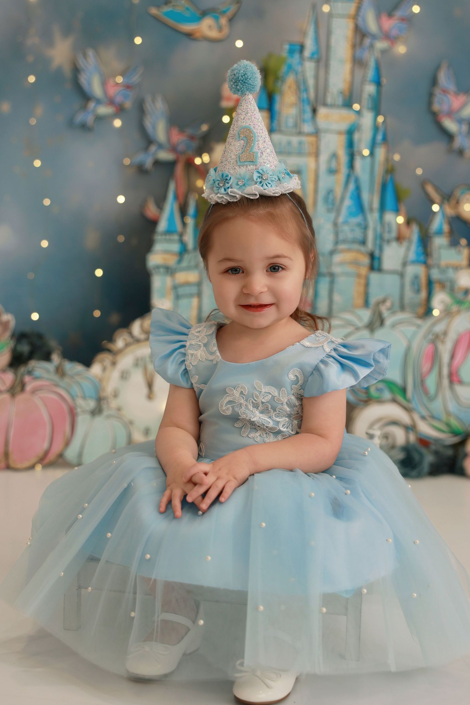 A baby in a light blue tulle party dress and silver crown poses against a magical winter wonderland backdrop with twinkle lights.