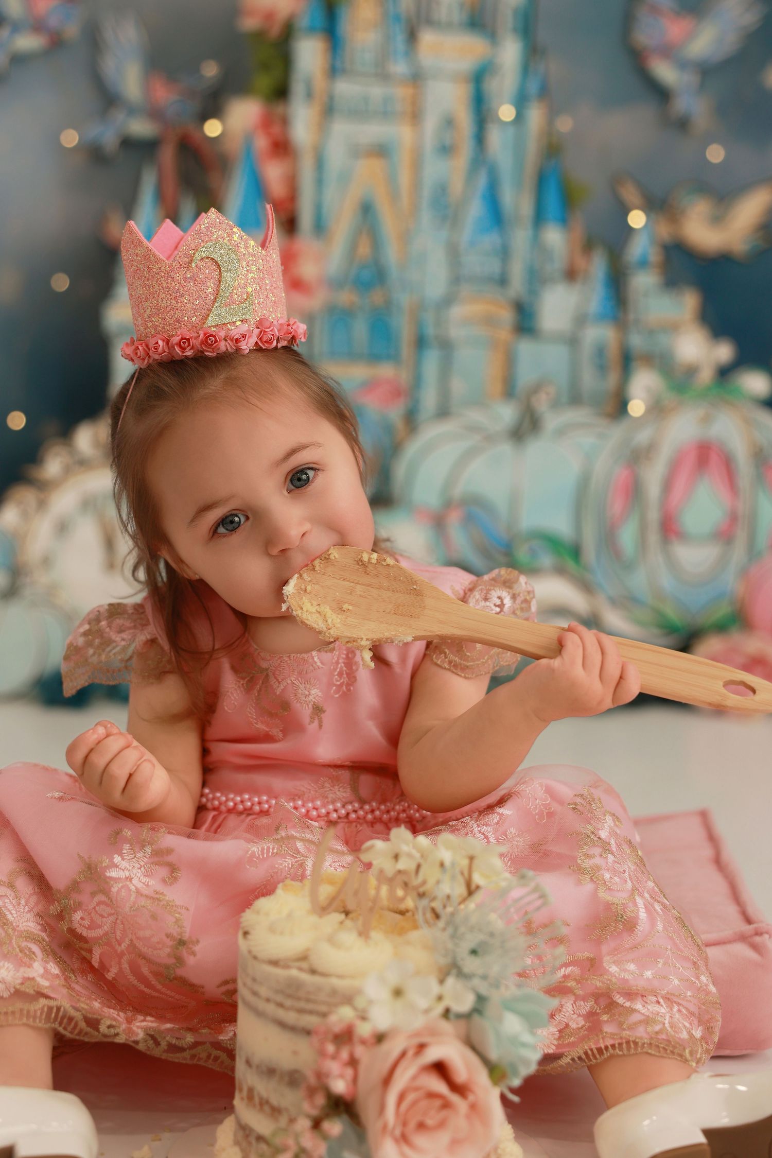 A baby in a pink dress and crown enjoys a cake smash photo session with pastel decorations and a castle backdrop.