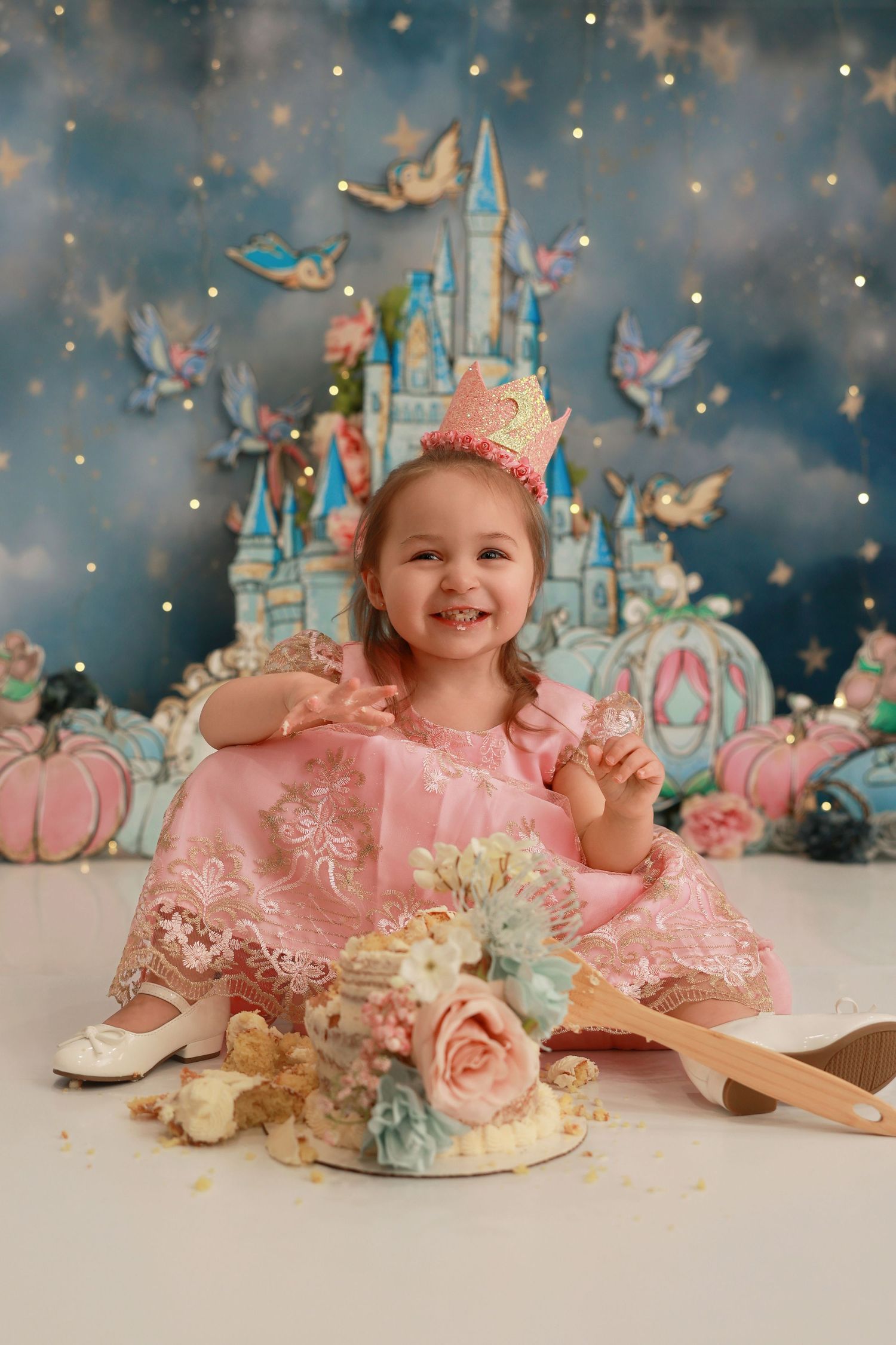 Magical princess themed photo setup with castle backdrop, butterflies, pink dress, and decorated cake against sparkly background.