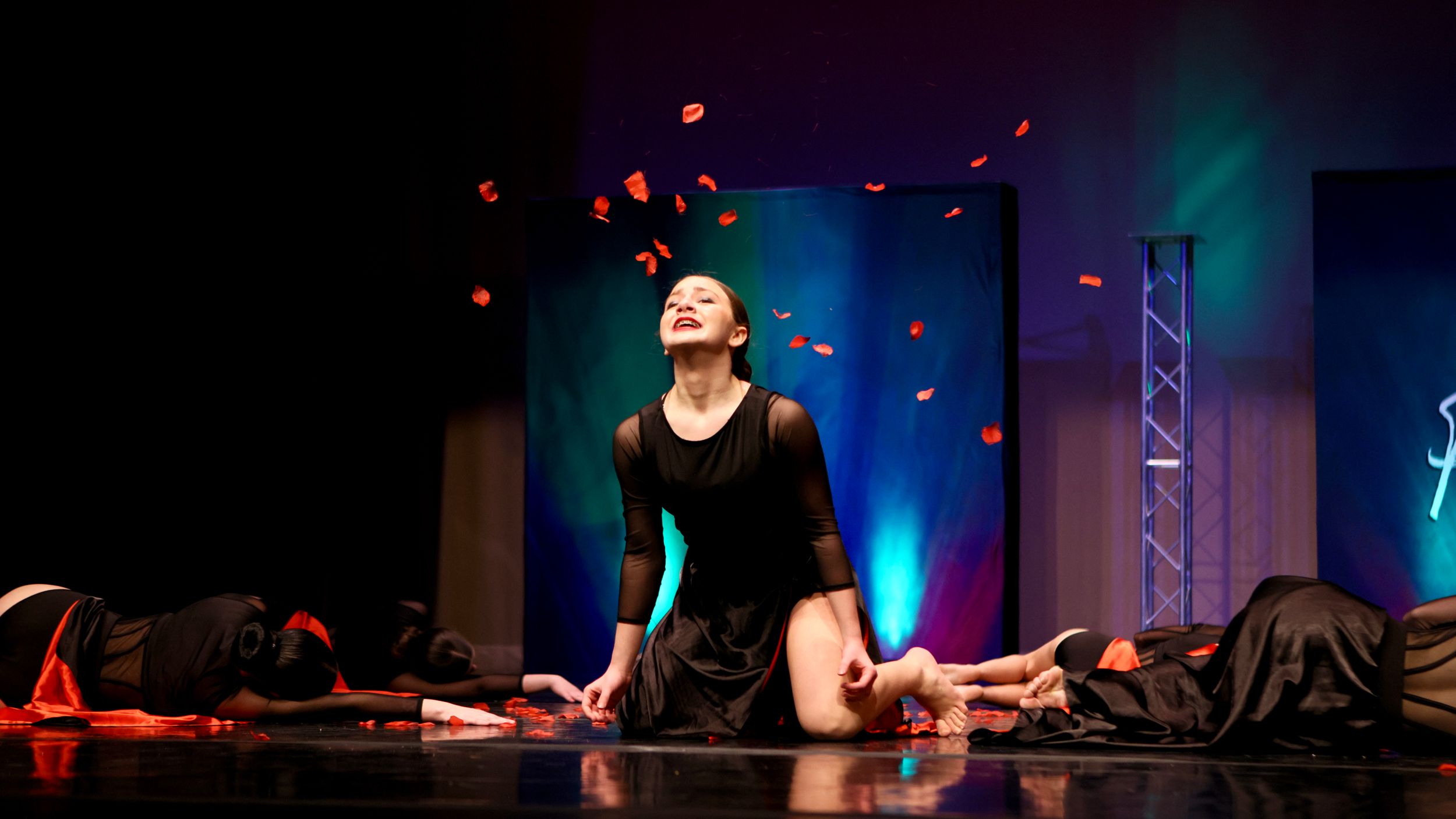 Female dancer in black dress performs emotional contemporary solo with falling petals on colorful stage.
