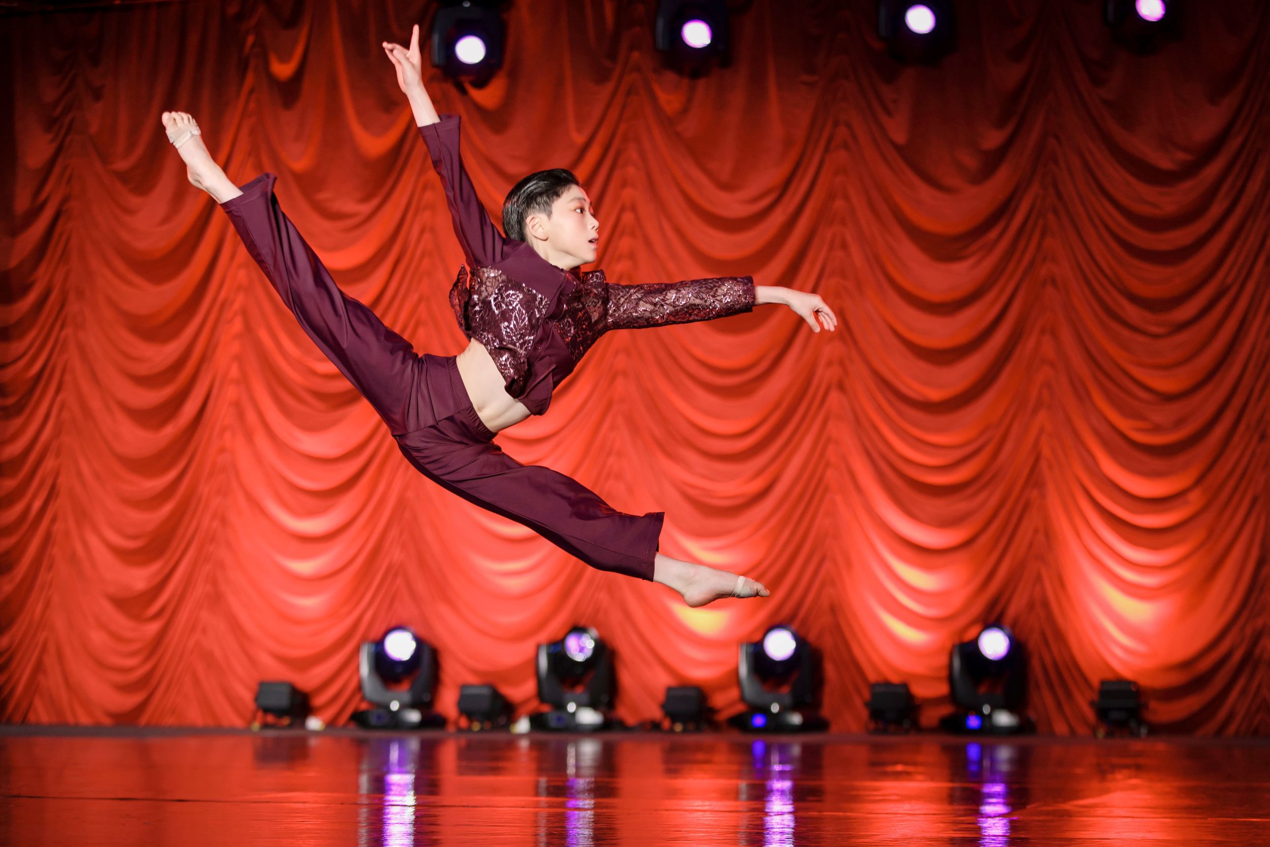 Male dancer in maroon outfit performs stunning aerial leap on stage with red curtain background.
