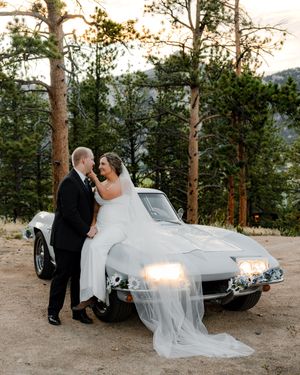 Bride and groom pose beside a vintage white Corvette surrounded by pine trees at a mountain wedding.