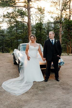 Bride and groom hold hands in front of a vintage car surrounded by pine trees at a Colorado mountain wedding.
