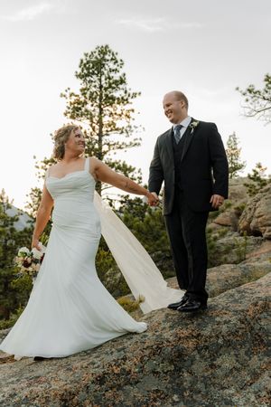 Bride and groom hold hands on rocky mountain overlook, she in white gown with veil, he in dark suit, pine trees behind.