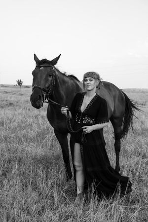 Black and white photo of a woman in a dark dress standing beside a horse in an open field.