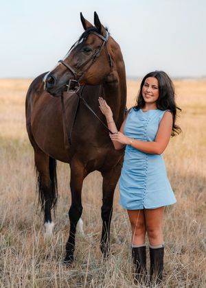 Young woman in blue dress and cowboy boots stands beside a bay horse in a golden field.