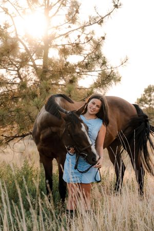 A smiling girl in a blue dress stands with a bay horse in a sunlit field with tall grass and pine trees.