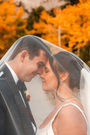 Bride and groom share an intimate moment under a veil with vibrant autumn foliage behind them.