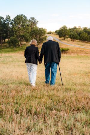 Elderly couple holding hands walking through a grassy field toward a road, man using a cane, both wearing black jackets.
