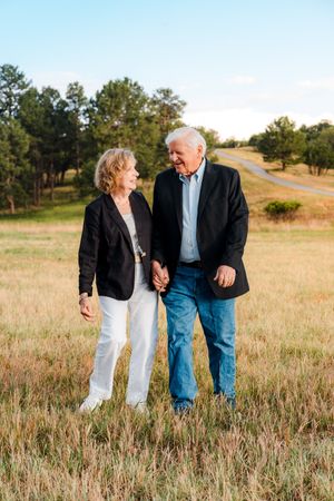 An elderly couple holds hands and smiles while walking through a golden grass field at sunset.