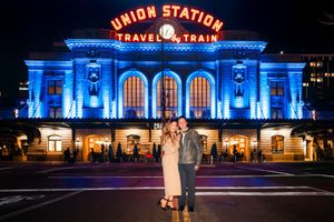 Couple embraces in front of Denver Union Station illuminated in blue lights at night.