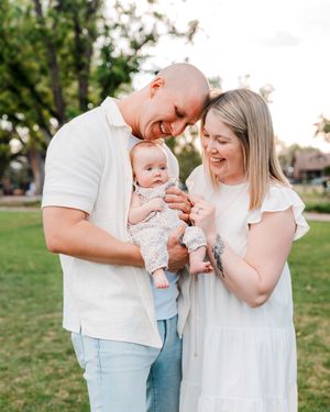 Happy family with bald father, blonde mother in white dress, and baby in floral outfit posing in a sunny green park.