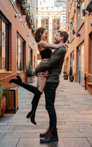 Man lifts smiling woman in a brick alley decorated with string lights in an urban setting.