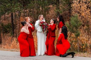 Bride in white gown poses with four bridesmaids in red dresses among autumn trees outdoors.