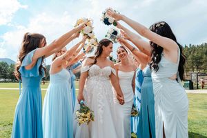 Two brides hold hands as bridesmaids in blue dresses raise bouquets overhead in an arch.
