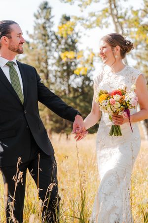 Bride and groom hold hands laughing in a sunlit golden meadow surrounded by autumn trees.
